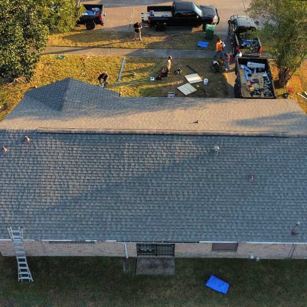 Overhead view of a house with roof replacement in progress; workers near trucks, debris containers, and a lawn.