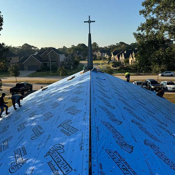 Church roof being shingled; workers on the blue-covered roof under a cross, with houses and cars in the background.