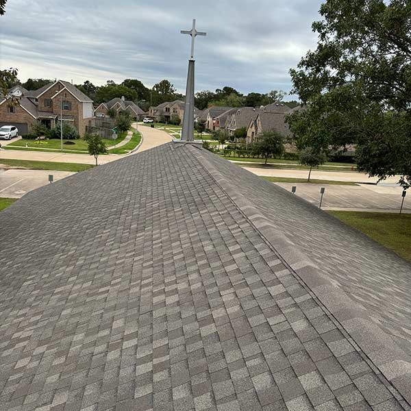 Gray shingle roof of a building with a cross-topped steeple. Suburban neighborhood in the background.