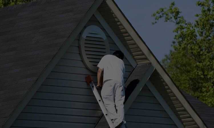 Person on a ladder painting the trim around an oval vent on a house with a gray roof.