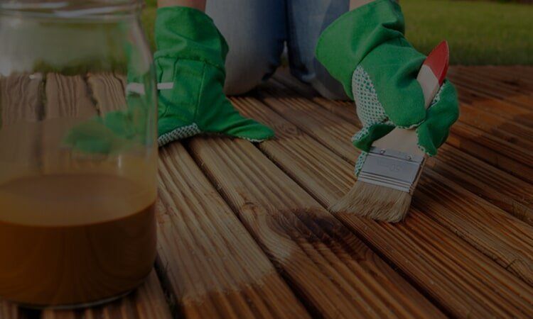 Person in green gloves staining a wooden deck with a brush and a jar of stain.