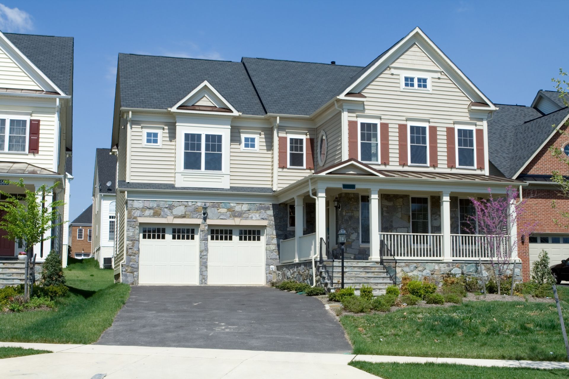 Two-story beige and gray house with a front porch, garage, and driveway on a sunny day.