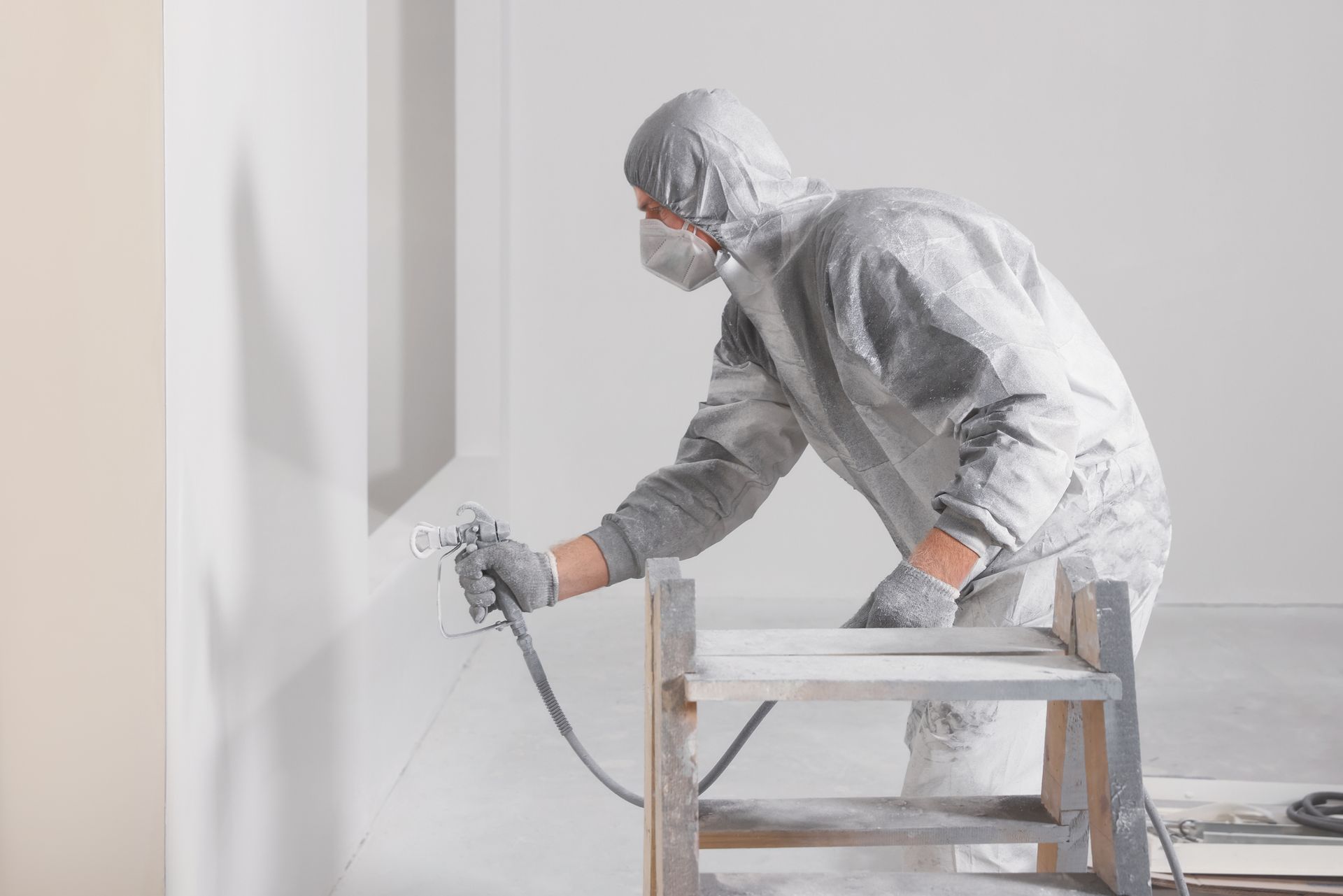 Man in protective suit spraying white paint on a wall using a sprayer.