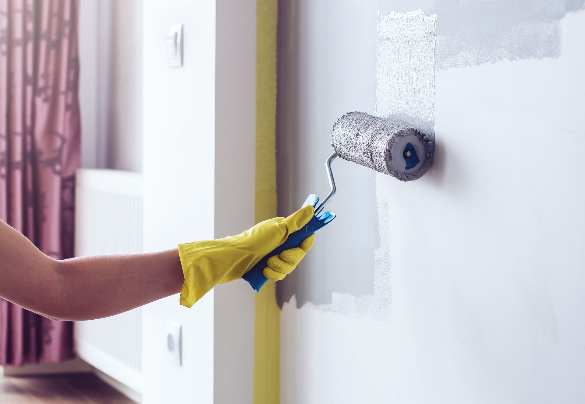Person wearing yellow gloves painting a white wall with a roller, interior setting.