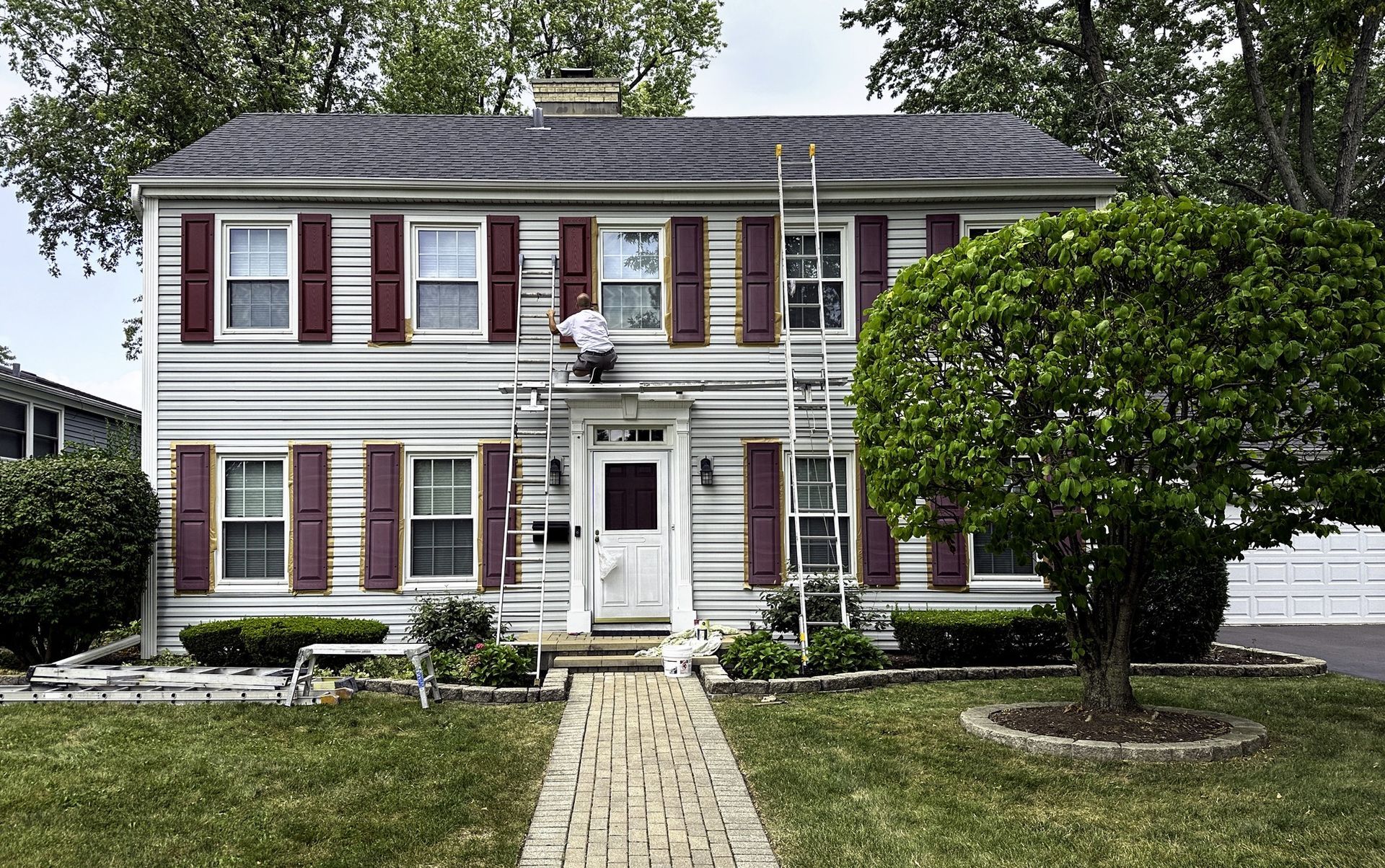 House with red shutters, a person on a ladder is working on it. Green tree and lawn.