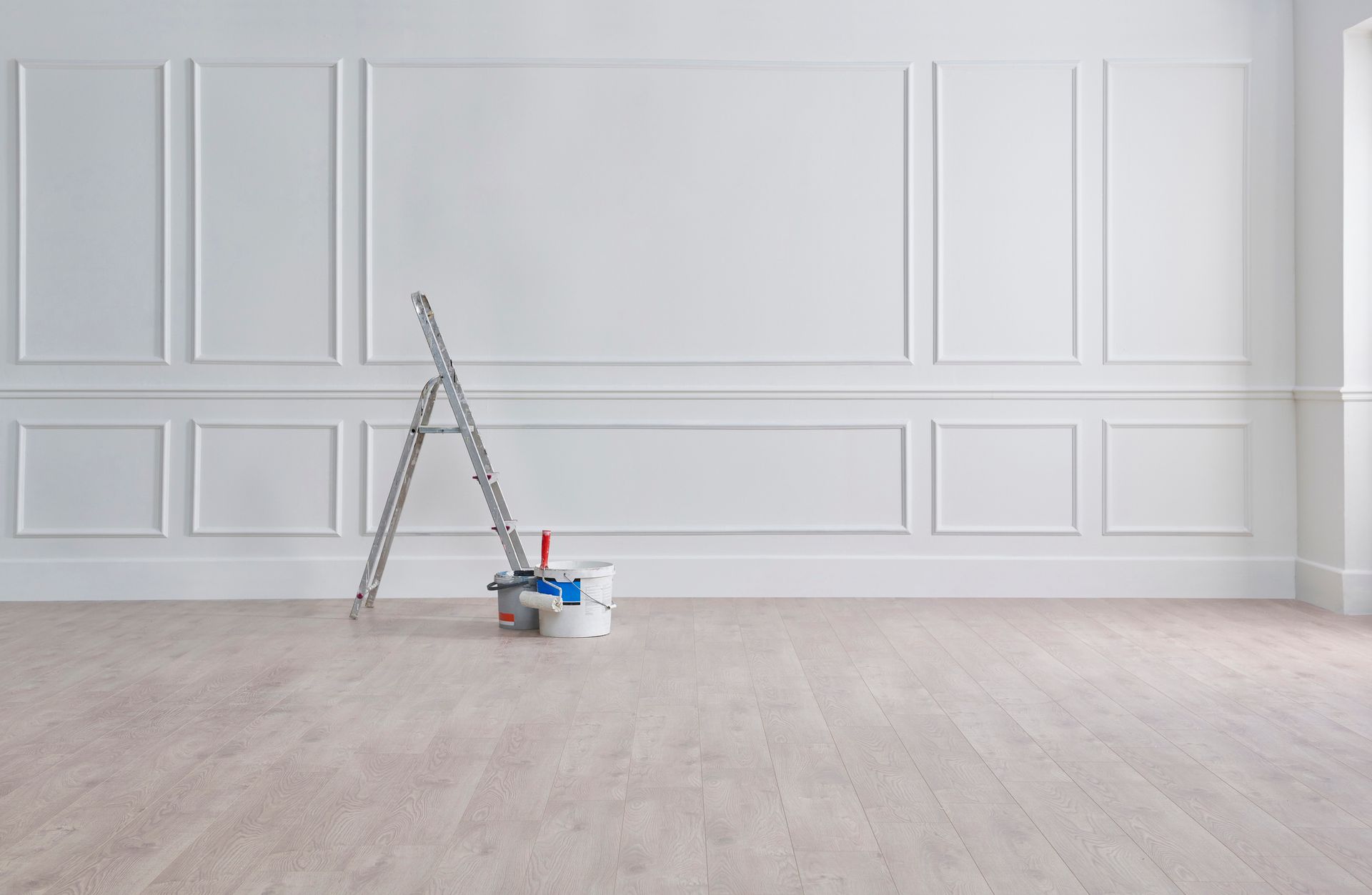 Ladder and paint supplies leaning against a white paneled wall, on a light wooden floor.