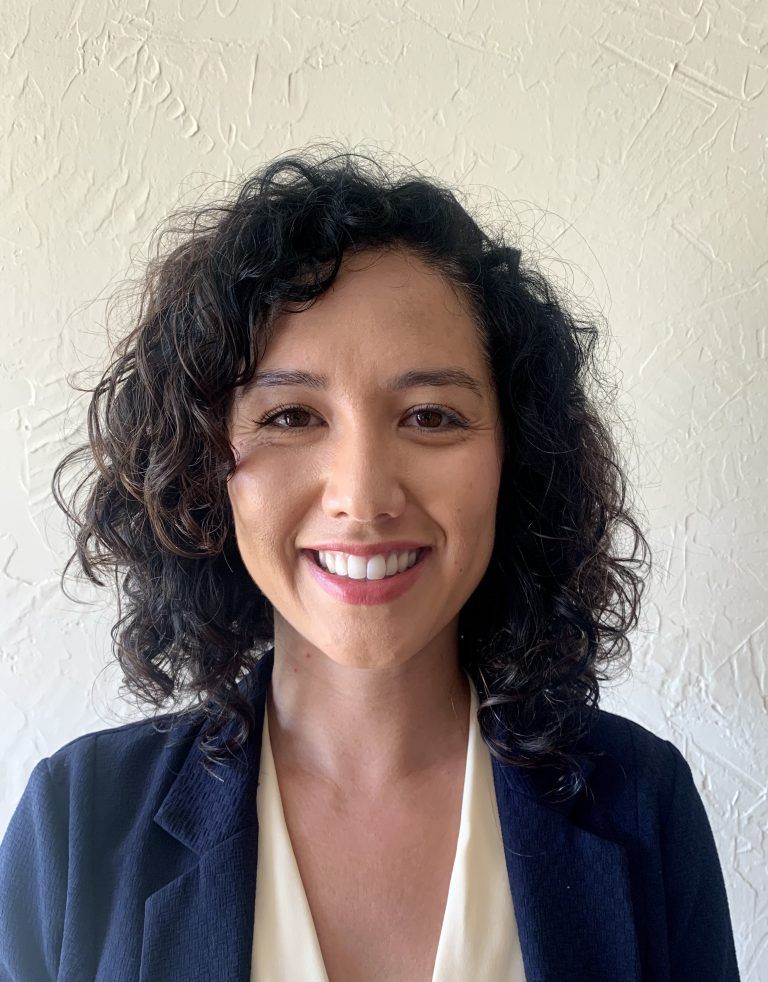 Woman with curly dark hair smiles, wearing a navy blazer over a light top, against a textured white wall.