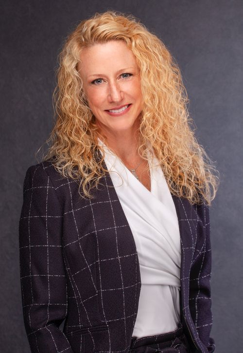 Woman with curly blonde hair smiles, wearing a white blouse and dark, plaid blazer.