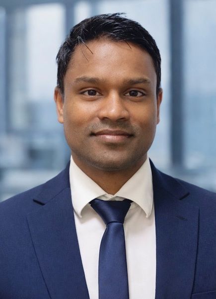 Man in a blue suit and tie, smiling at the camera, against a gray background.