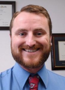 Man with red beard, blue shirt, red tie, smiling in an office setting.