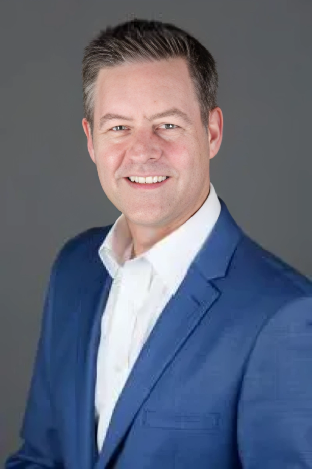 Man in blue suit and white shirt smiles at the camera against a gray background.