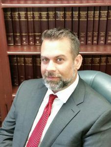 Man in suit and red tie sits in a chair in front of a bookcase.