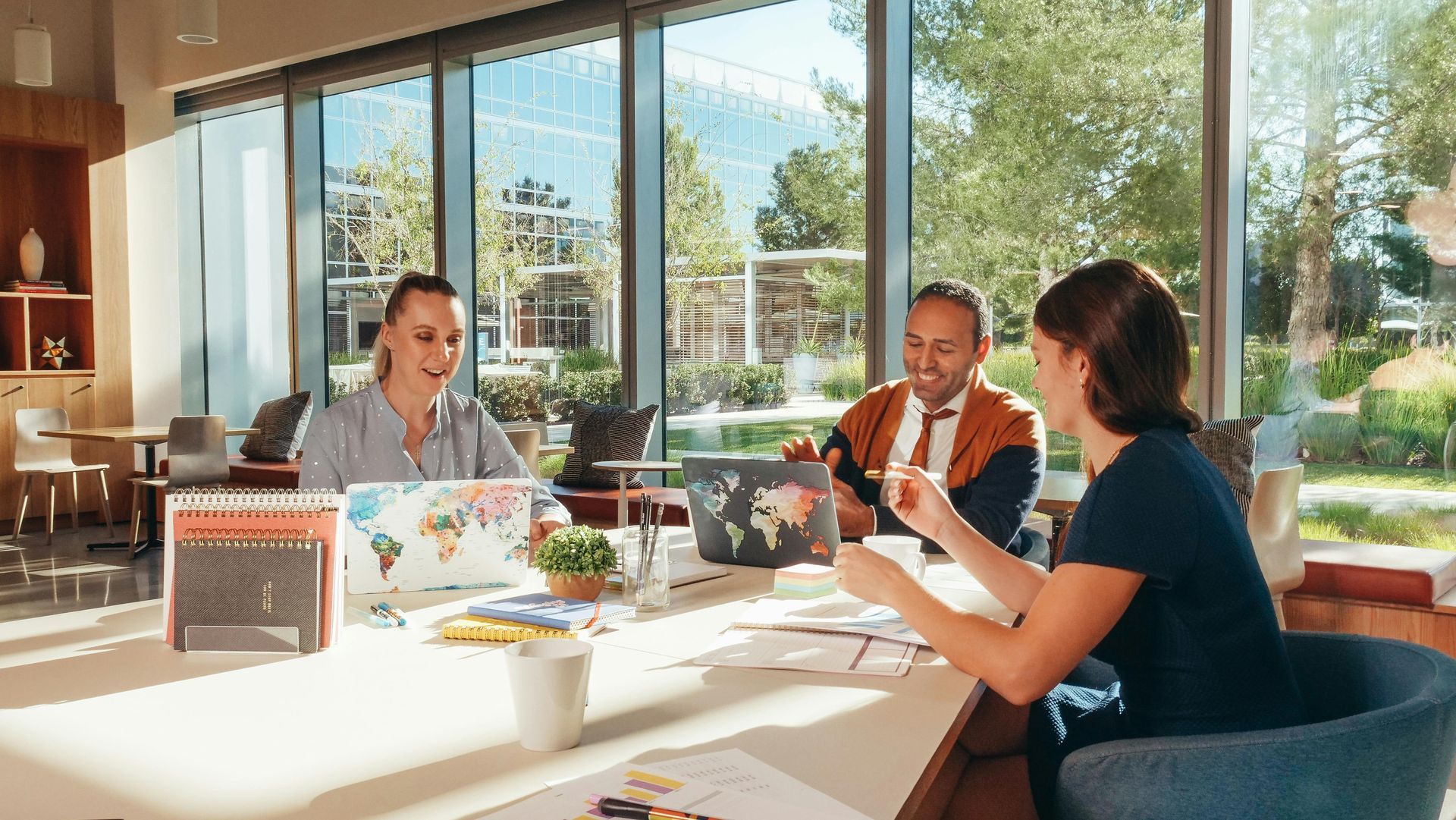 Three people collaborate around a table with laptops near large windows.
