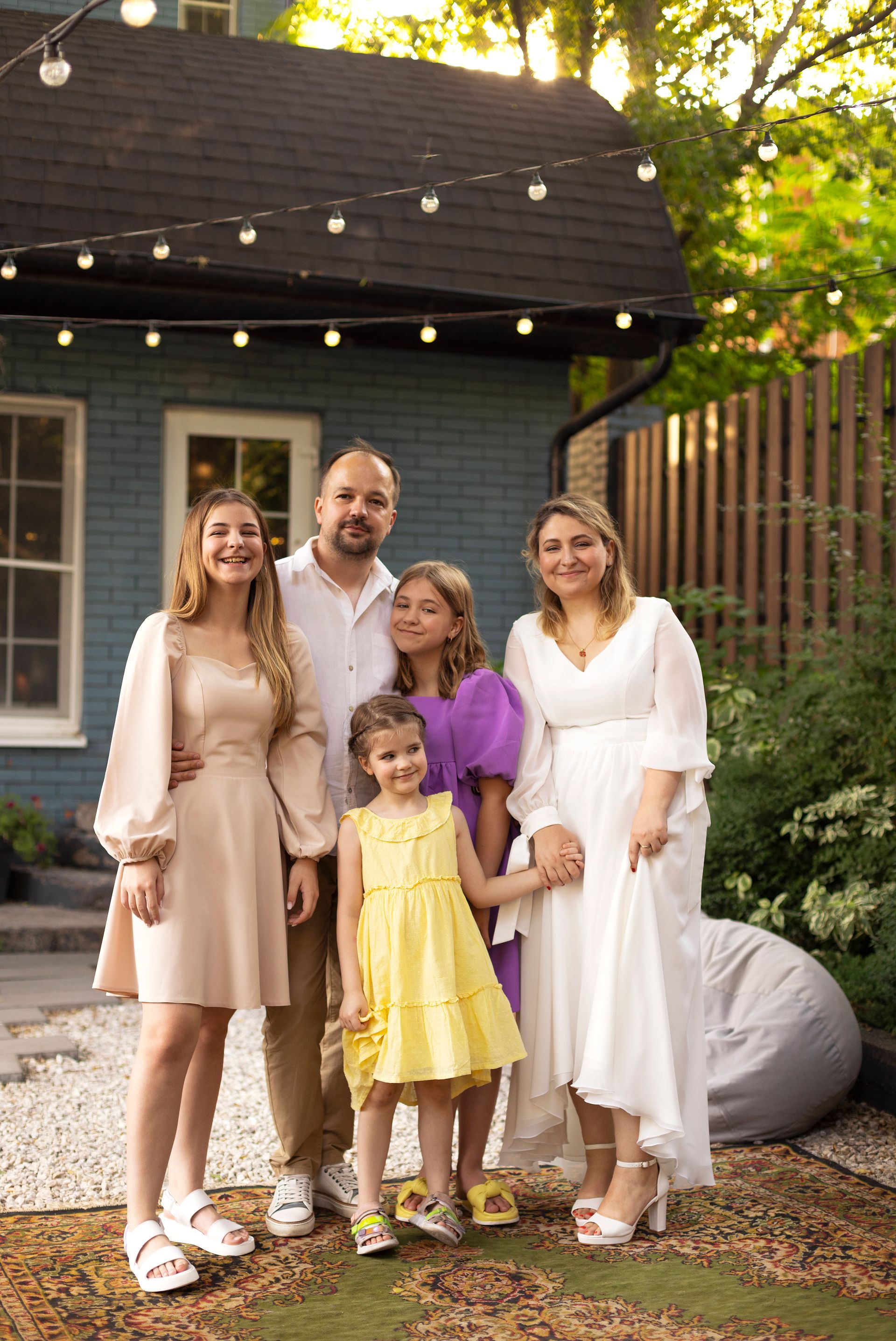 Family of five posing outdoors in front of a blue house, smiling.