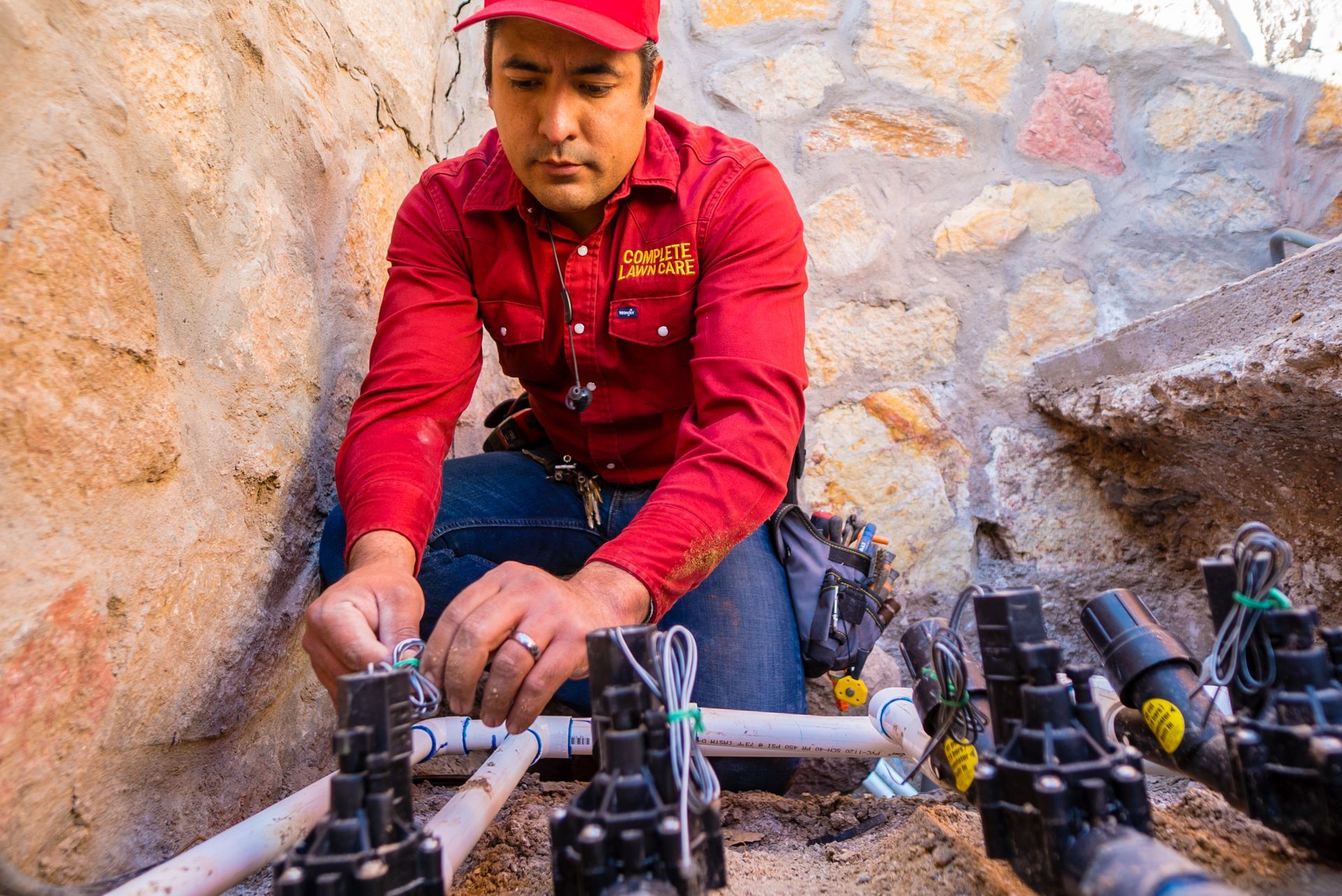 A professional on sprinkler system technician in a red shirt and hat is working on a pipe.