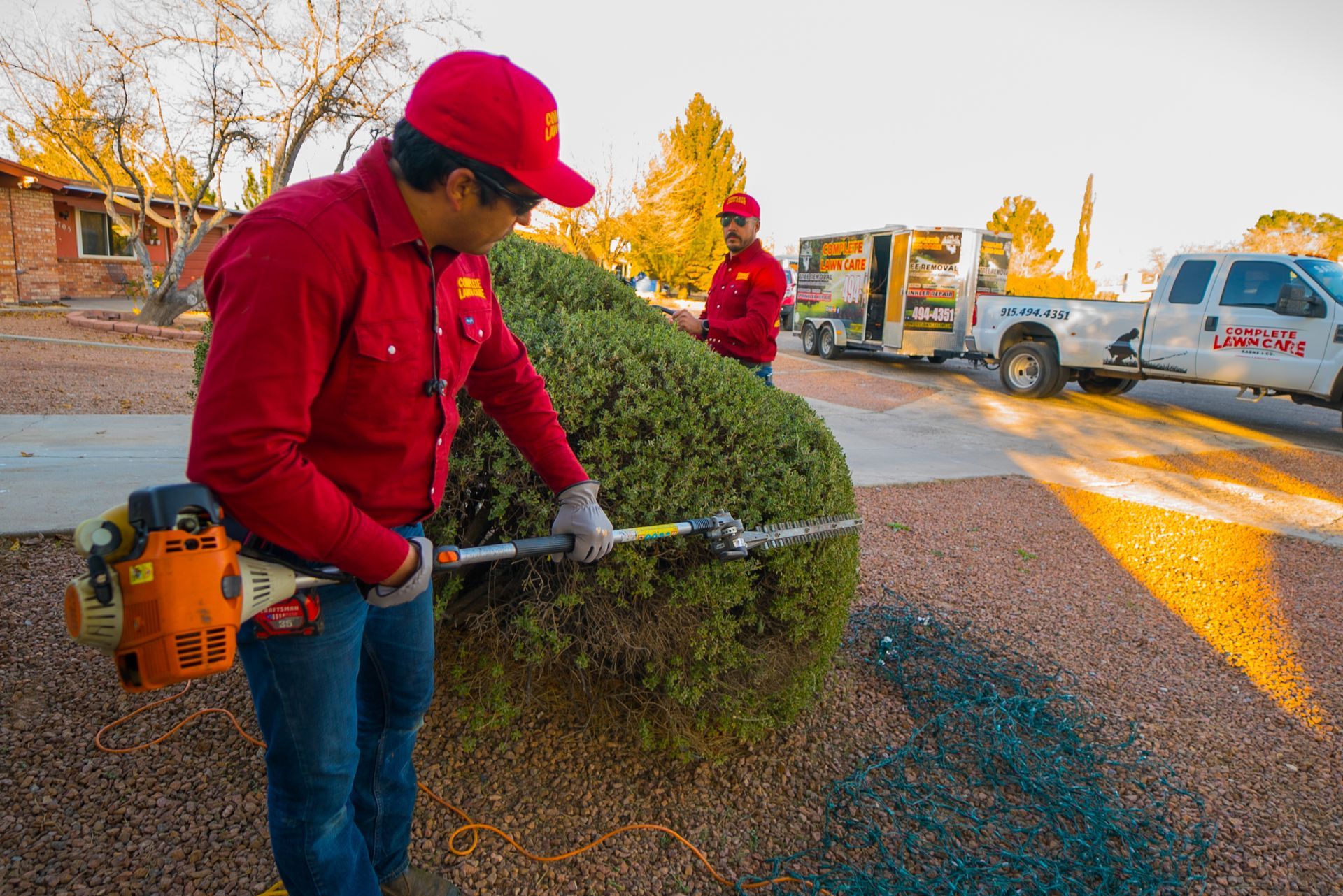 Two professional landscapers from Complete Lawn Care in red uniforms are pruning a large bush with precision tools. A Complete Lawn Care truck and trailer are parked in the background, showcasing their equipment and branding. The setting captures the warm light of sunset.