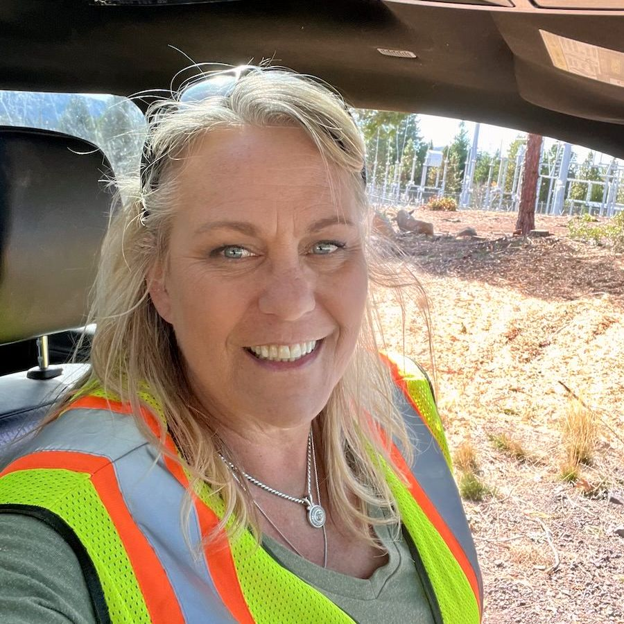 Woman in a safety vest smiles in a vehicle near utility poles.