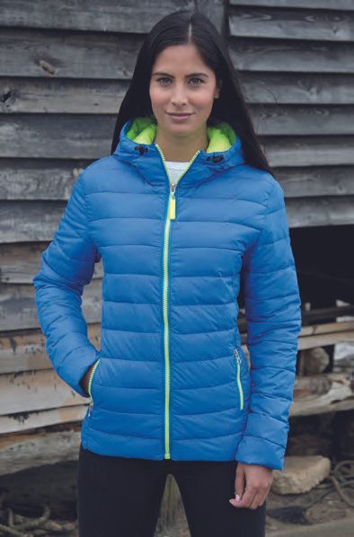 Μια γυναίκα που φοράει μπλε μπουφάν με κουκούλα στέκεται μπροστά σε ένα ξύλινο κτίριο.