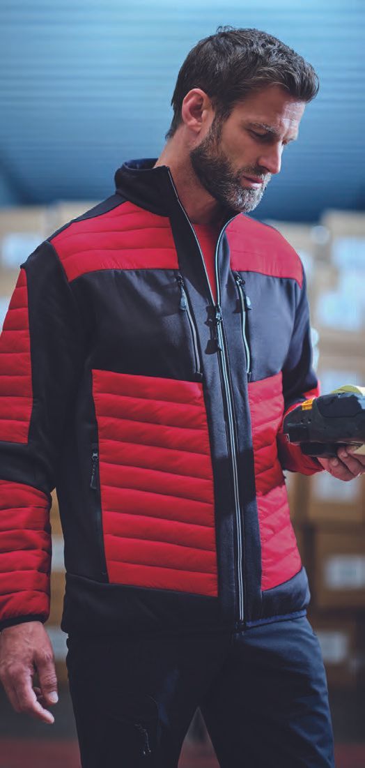 Ένας άντρας που φοράει κόκκινο και μαύρο μπουφάν στέκεται σε μια αποθήκη κρατώντας ένα κινητό τηλέφωνο.