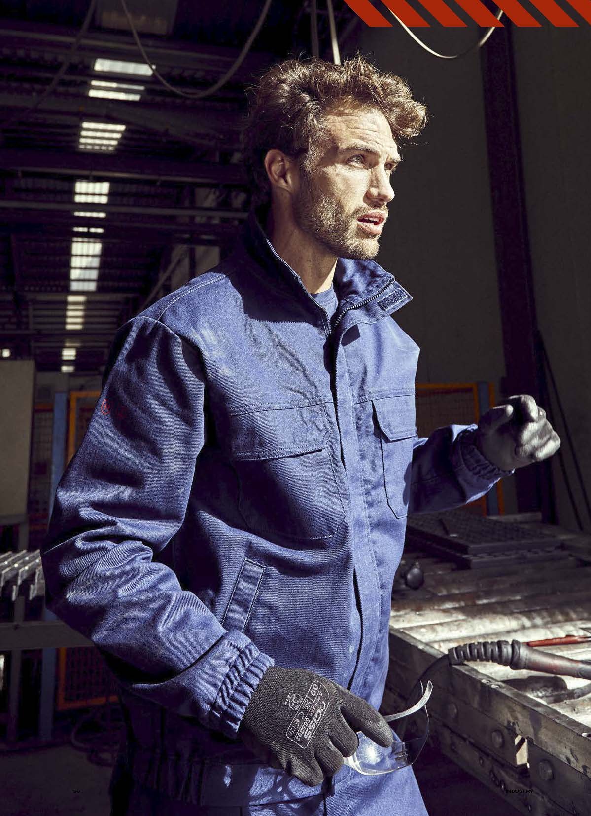 Ένας άντρας που φοράει μπλε μπουφάν και μαύρα γάντια στέκεται σε ένα εργοστάσιο.