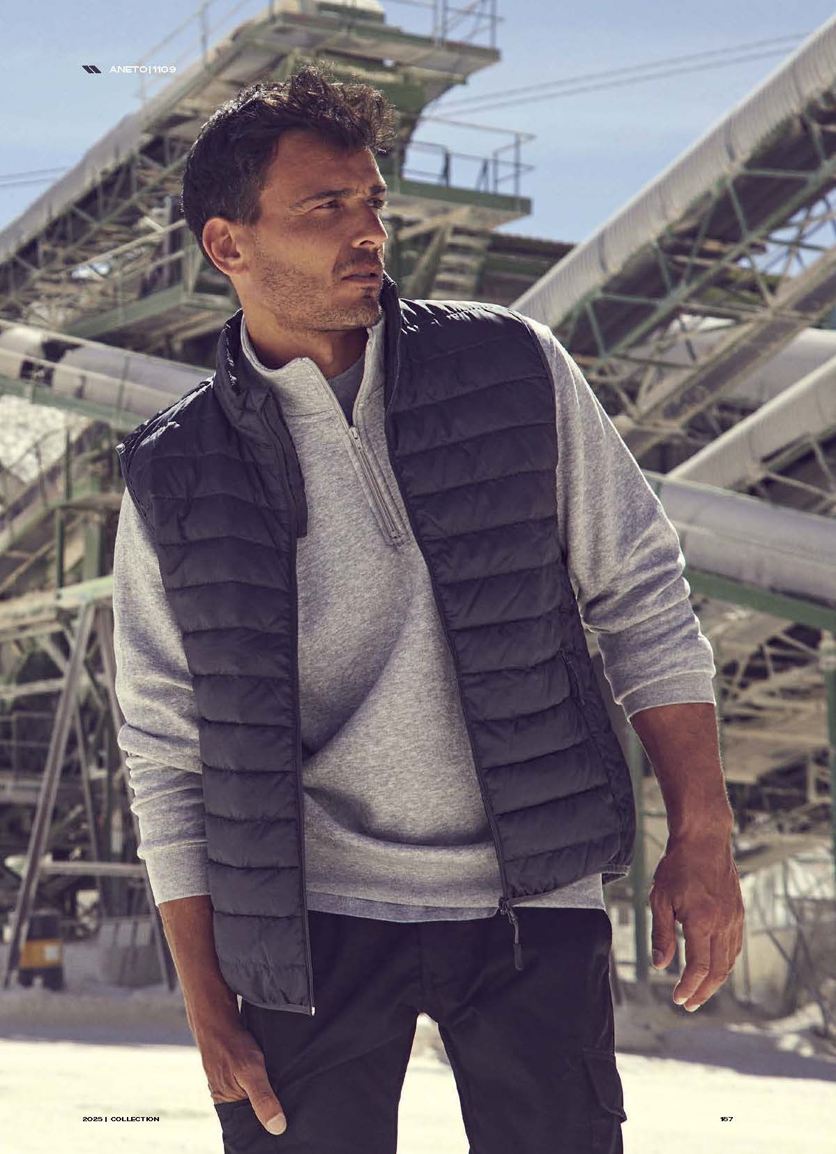 Ένας άντρας που φοράει γιλέκο και πουλόβερ στέκεται μπροστά σε ένα κτίριο.