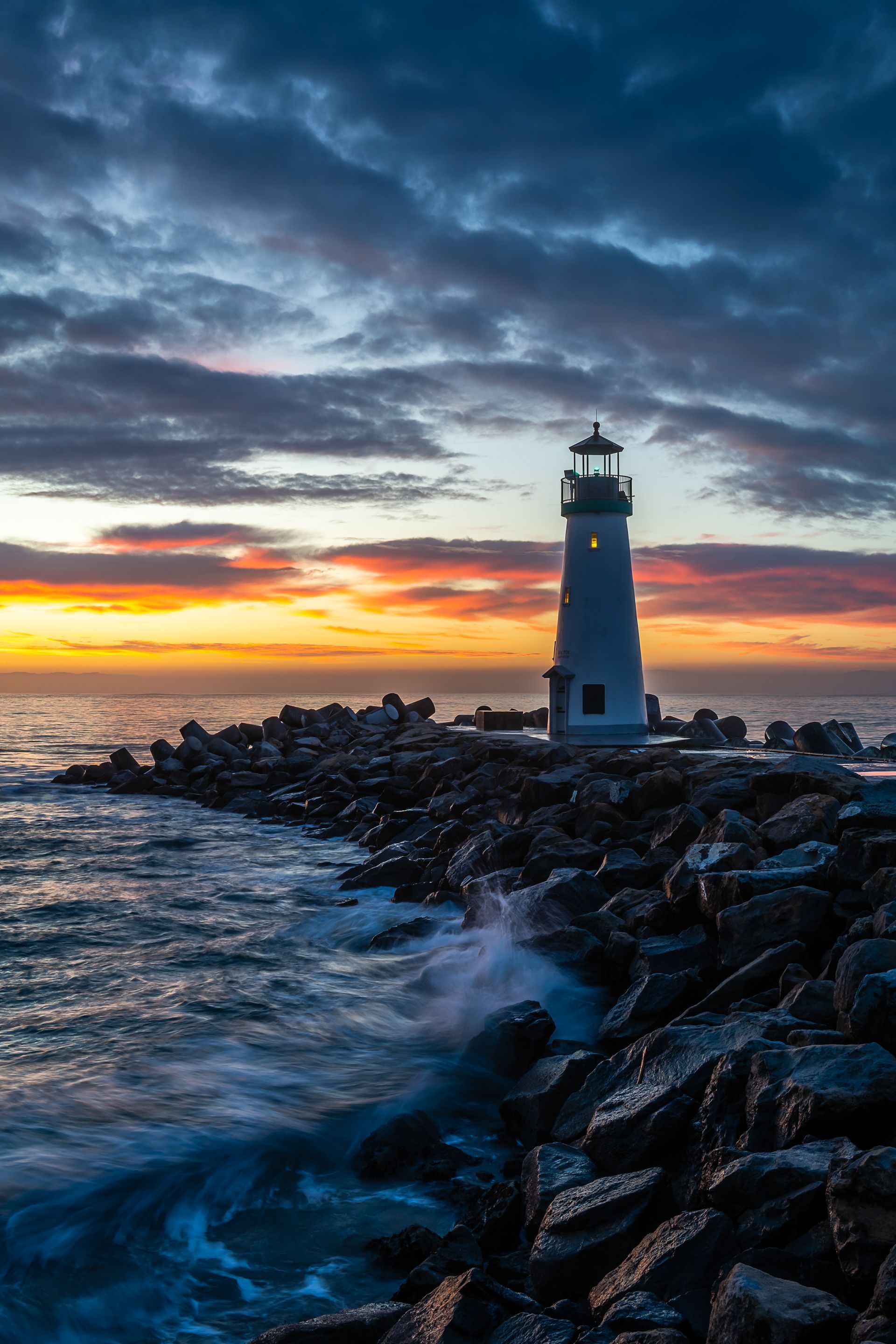 A lighthouse is sitting on a rocky shoreline next to the ocean at sunset.