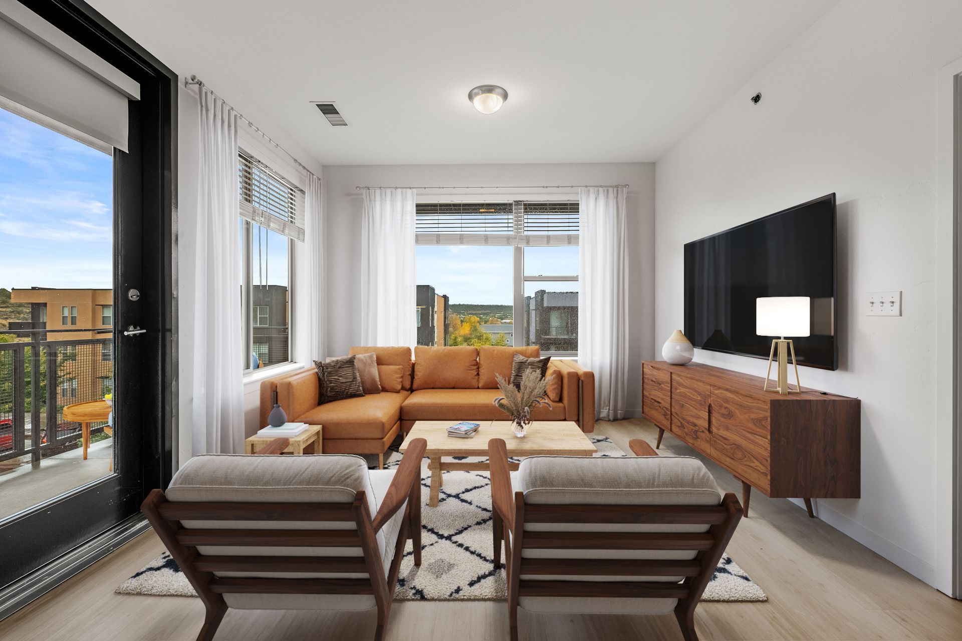 Bright apartment living room with tan sectional sofa, wooden coffee table, and a wall-mounted TV.