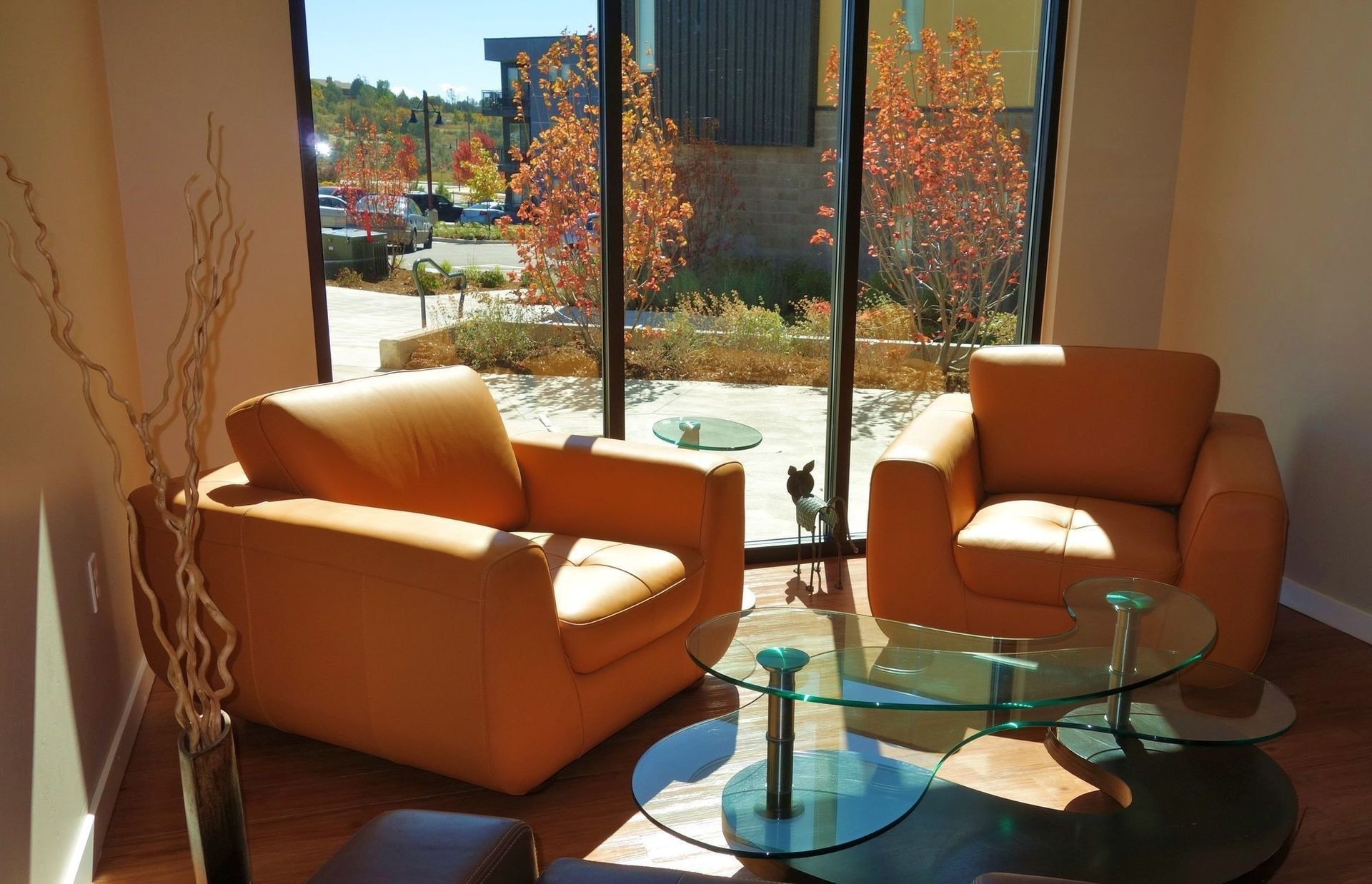 Interior community lounge with two orange leather chairs and glass coffee tables by a large window.