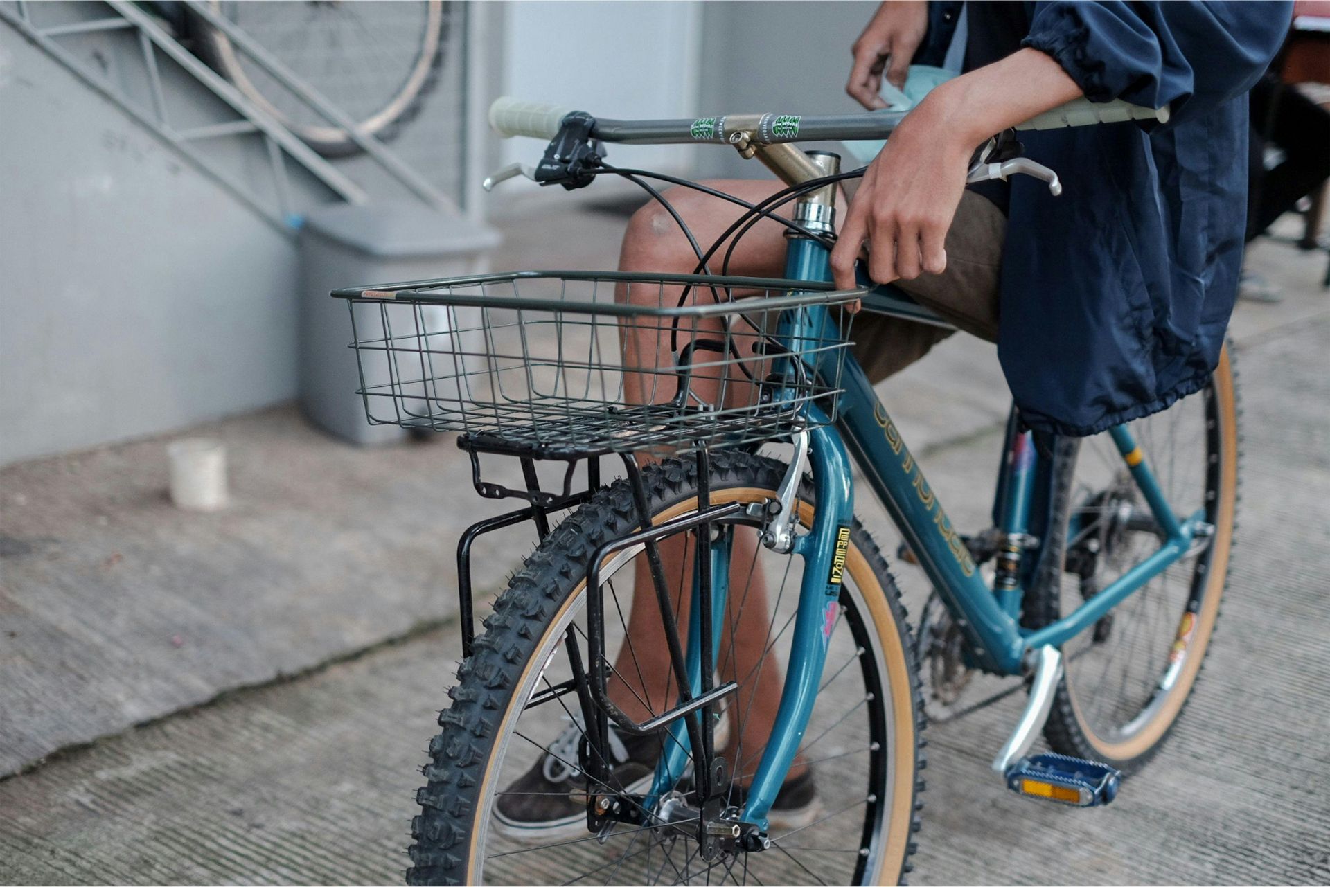 A close-up of a person sitting on a teal bicycle with a wire basket mounted on the front handlebars.