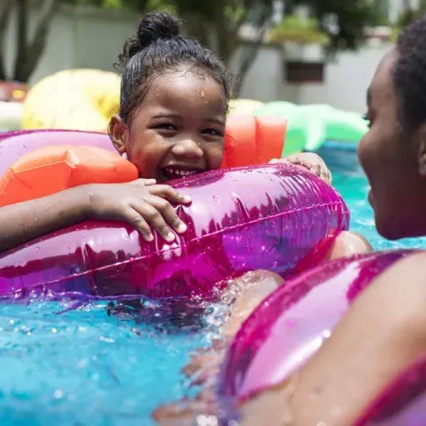 A person and a child in a swimming pool, both using pink inflatable rings and orange arm floaties, smiling at each other.