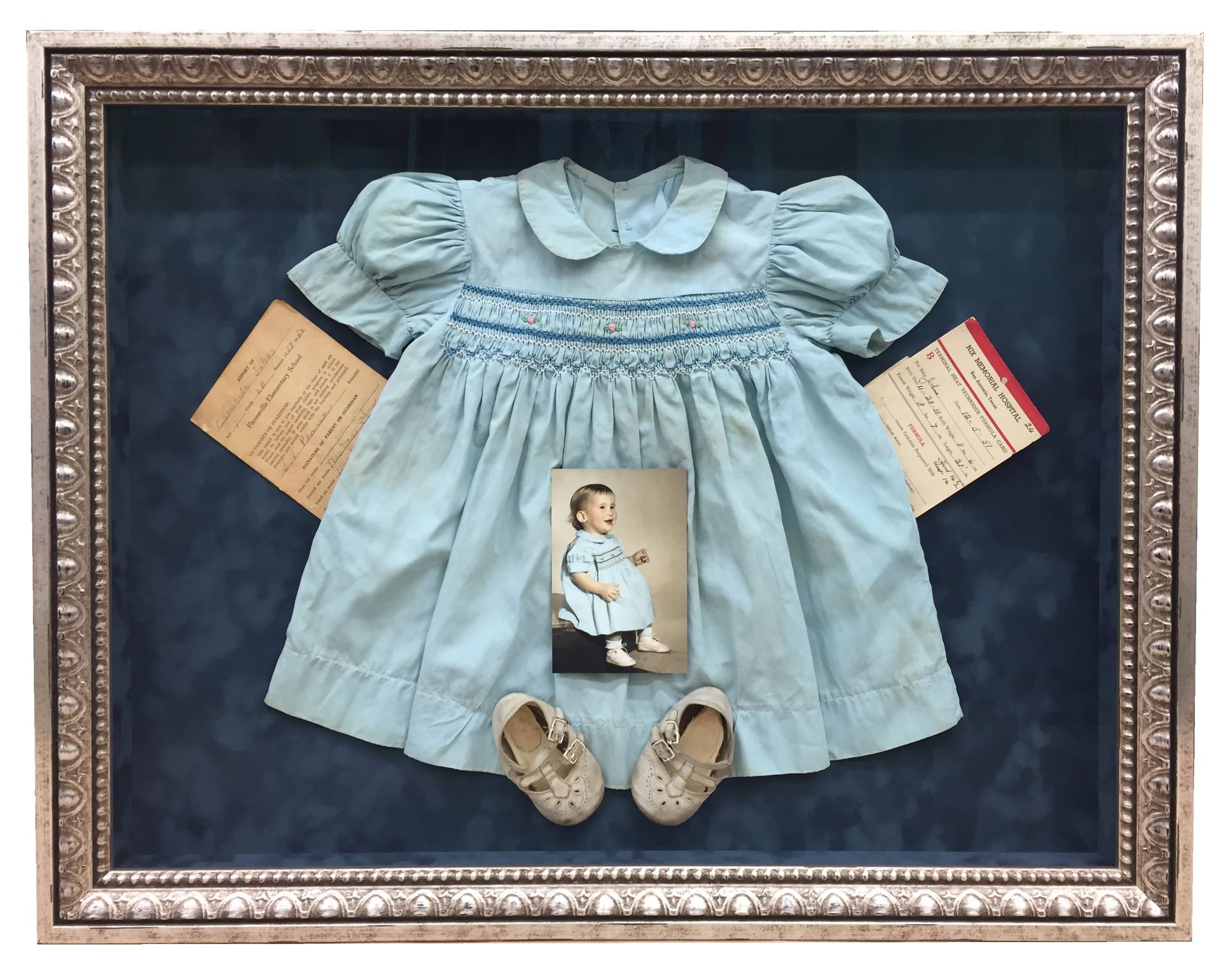 A framed display with a blue dress, baby shoes, photo, and papers.