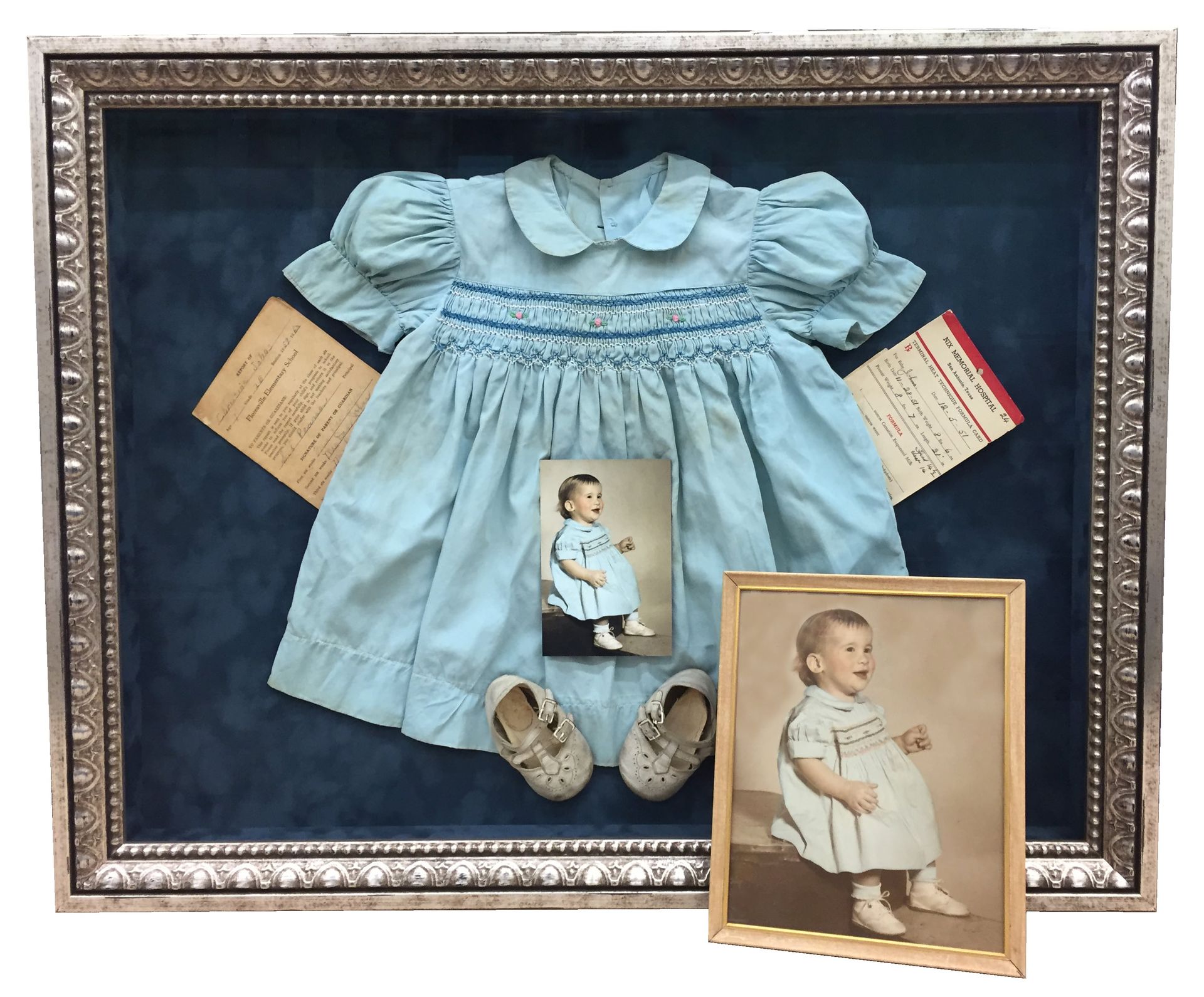 Shadowbox with a baby dress, shoes, photos, and documents, all in shades of blue and sepia.