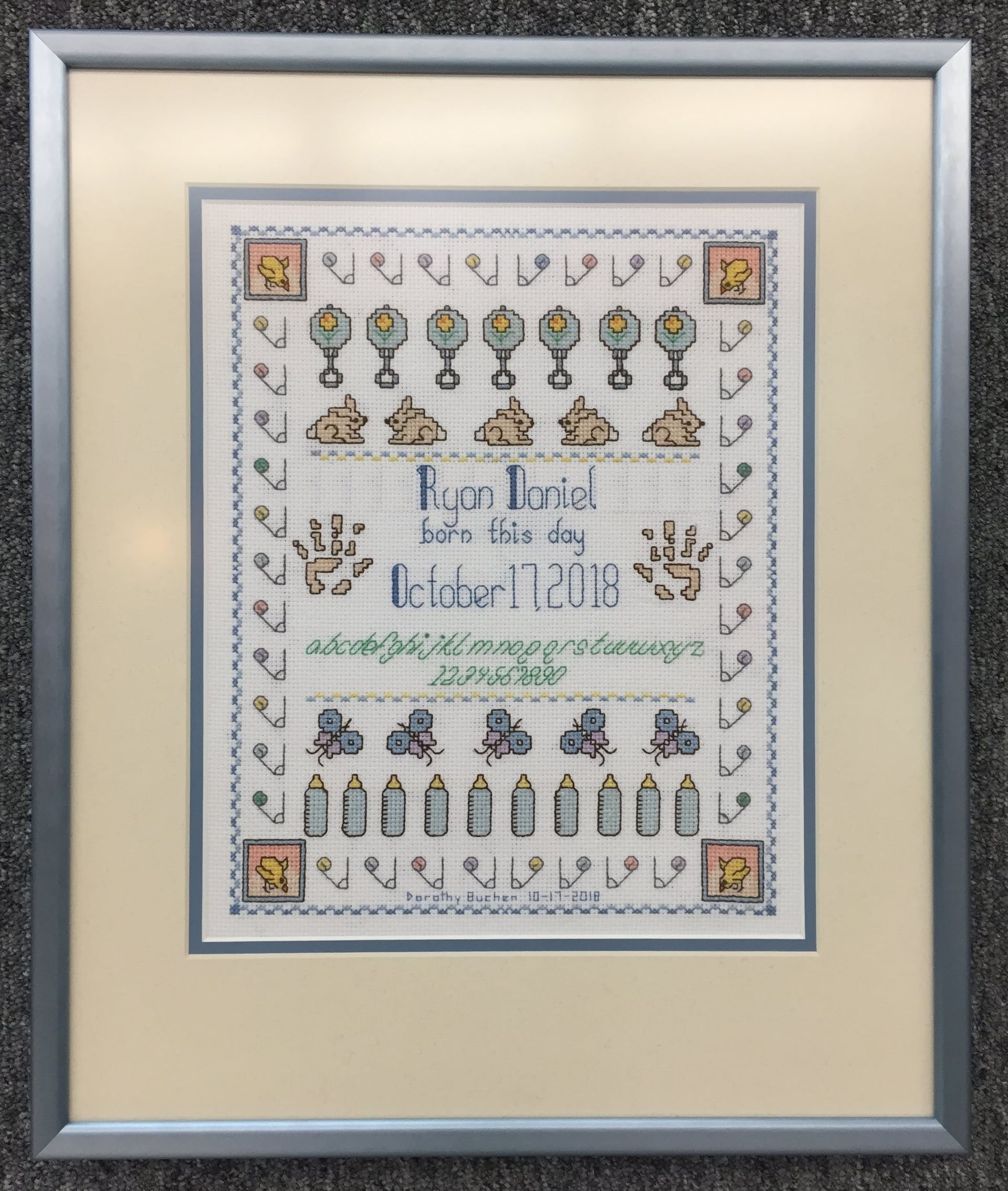 Framed cross-stitch baby announcement with birth date, name, and decorative elements in a silver frame.