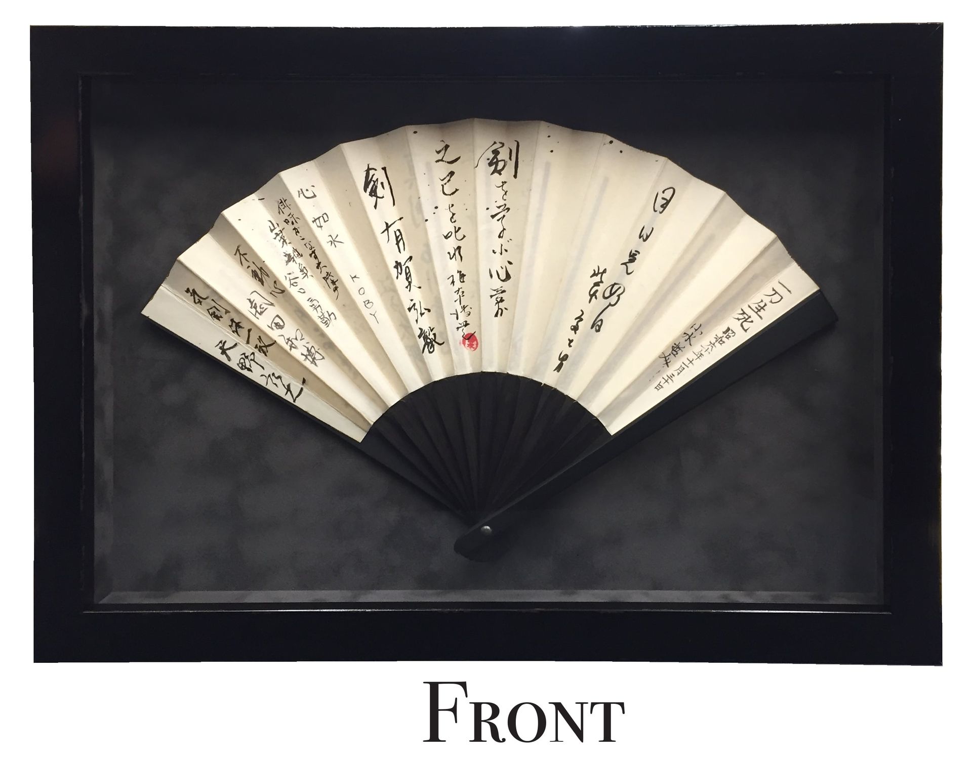 A framed folding fan with black calligraphy on white paper and a black handle against a dark gray background.