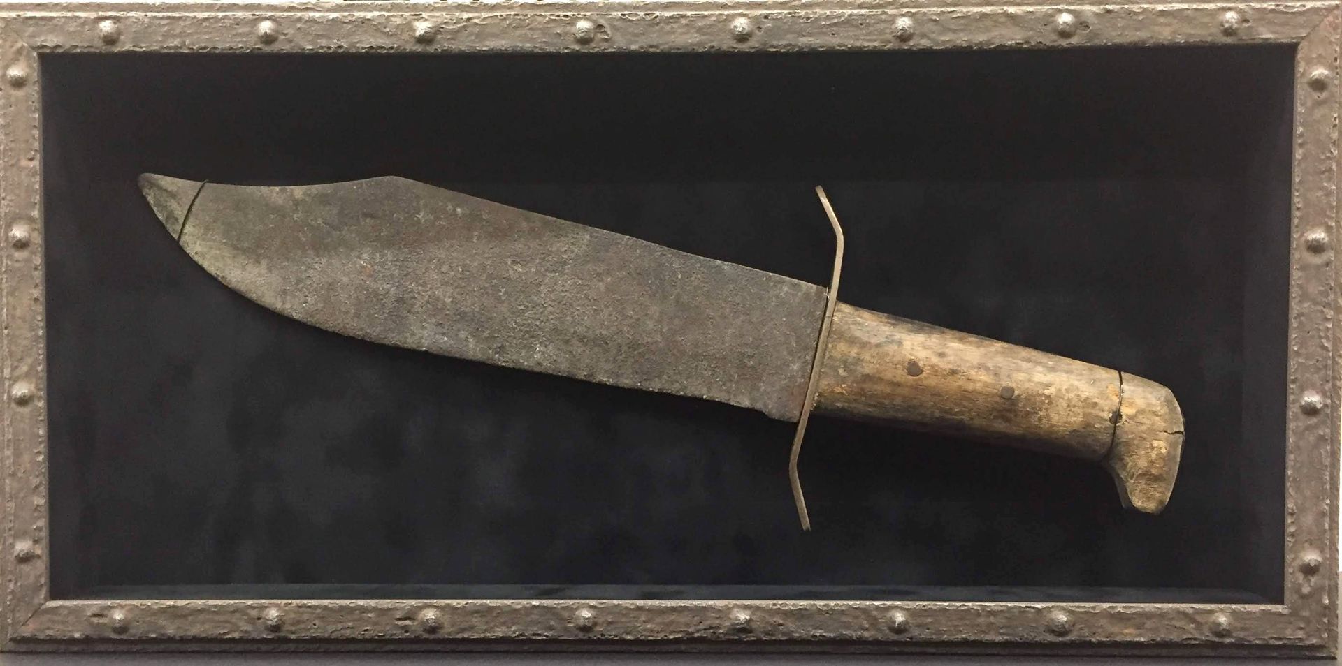 A rusty bowie knife in a shadow box with a dark background and metal frame.