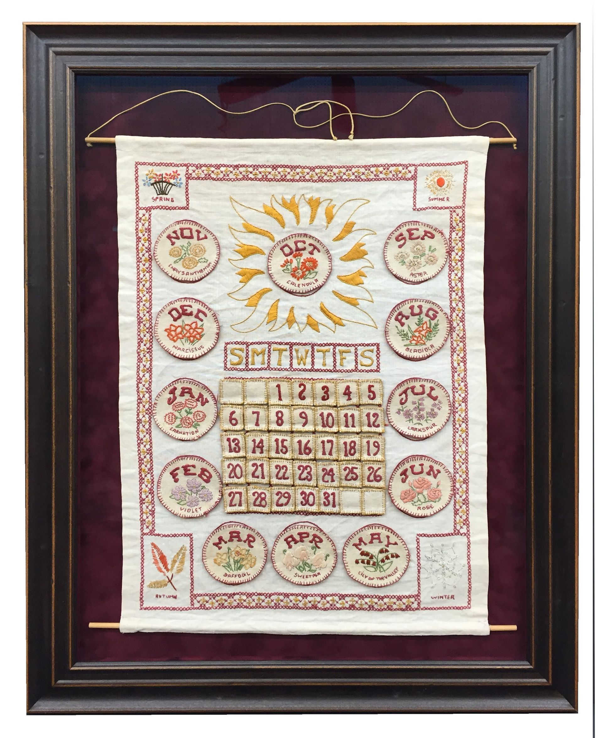 Framed embroidered calendar with floral motifs and dates; displayed with hanging wire.
