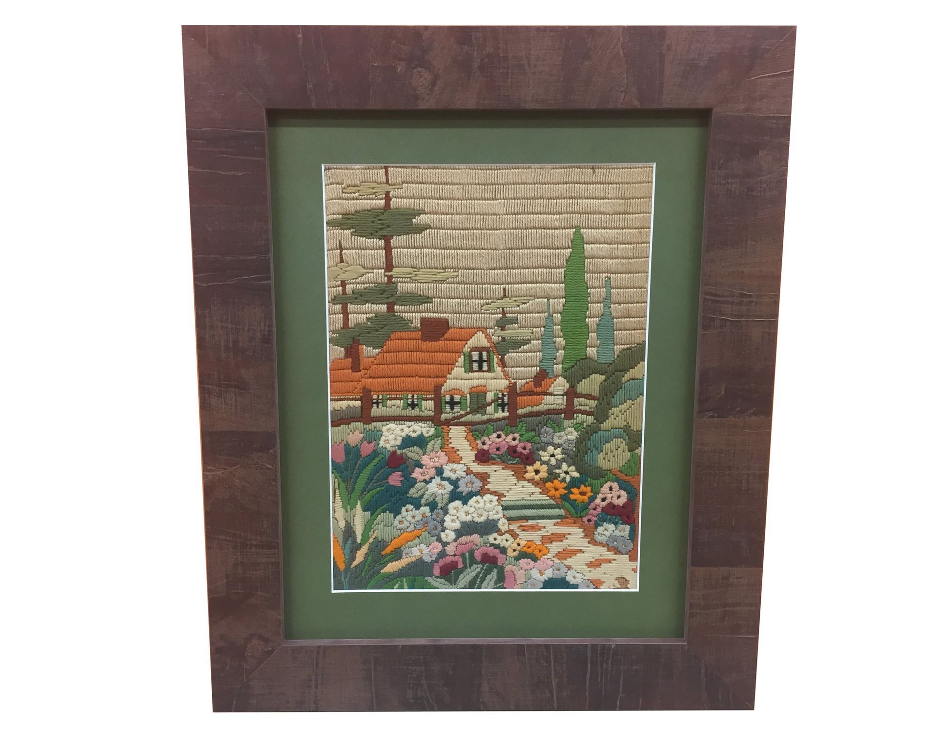 Needlepoint of a house and garden framed in brown. Green mat, flowers and trees are in the garden.