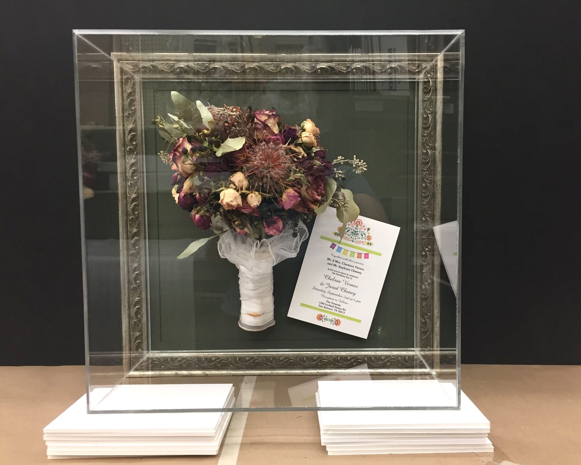 A framed dried flower bouquet with a note inside a clear acrylic display case.