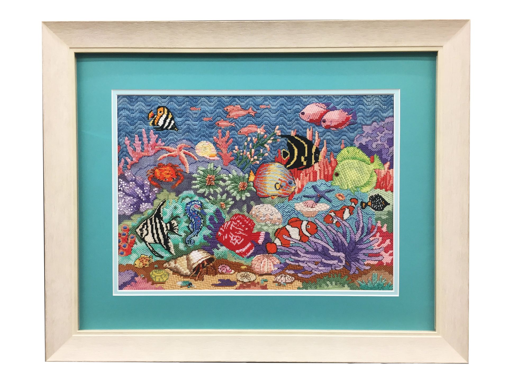 Framed cross-stitch art of a colorful coral reef with various fish in a light blue mat and cream frame.