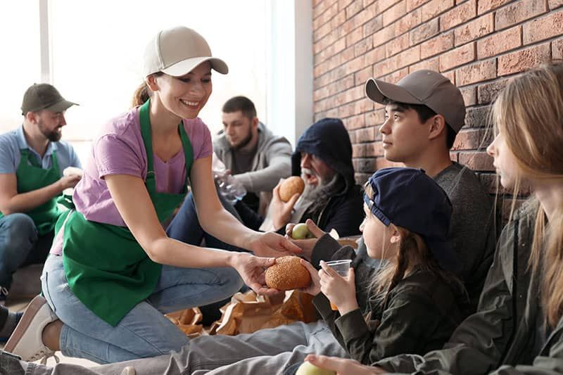 A young woman offering food to people sitting outside against a brick wall