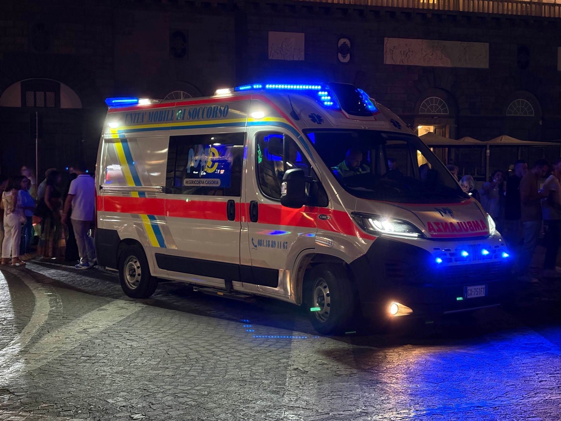 Ambulanza con luci blu lampeggianti parcheggiata di notte, persone nelle vicinanze.