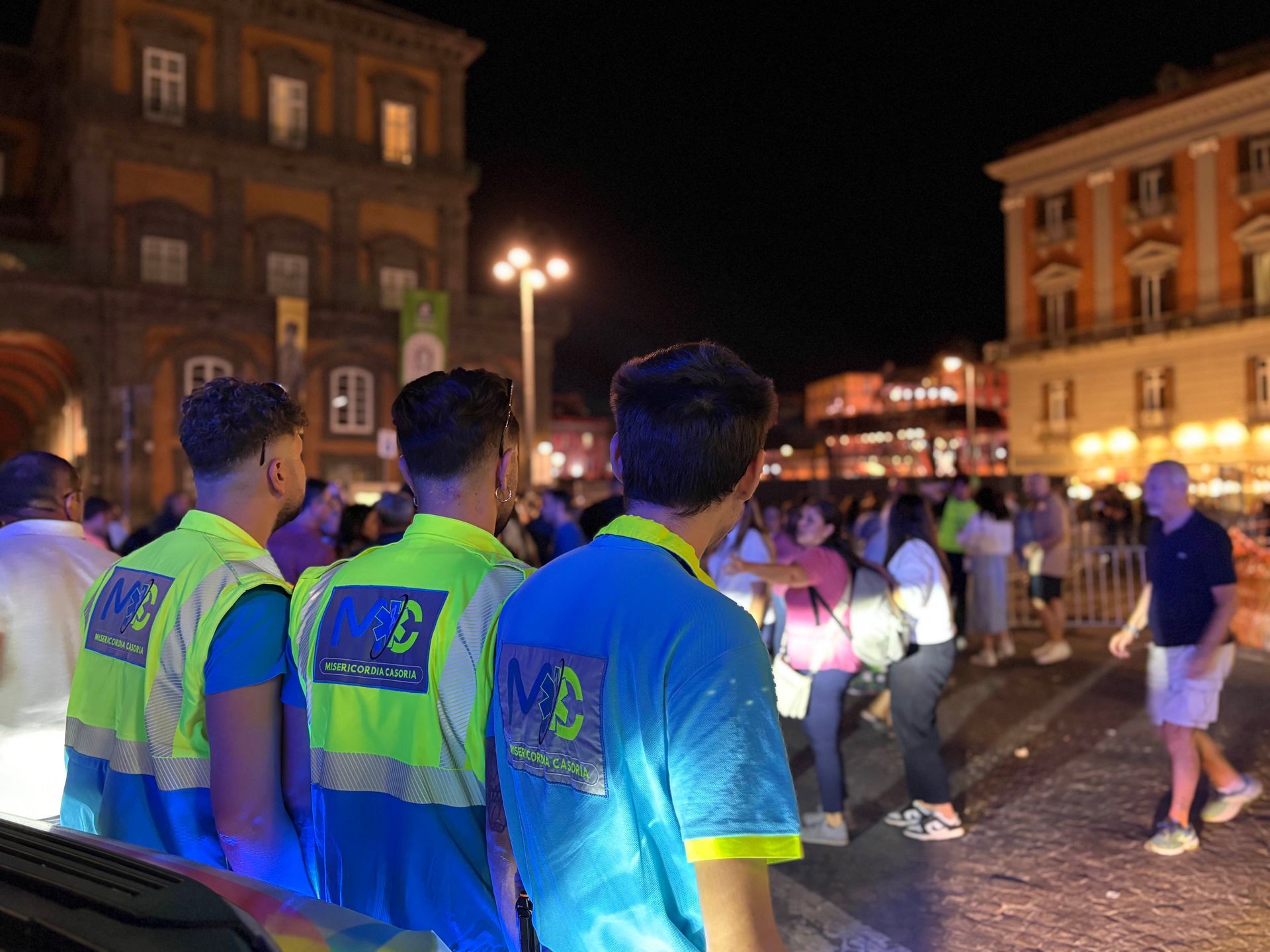 Tre persone con gilet color neon e camicie blu, di fronte alla folla di notte, con edifici sullo sfondo.