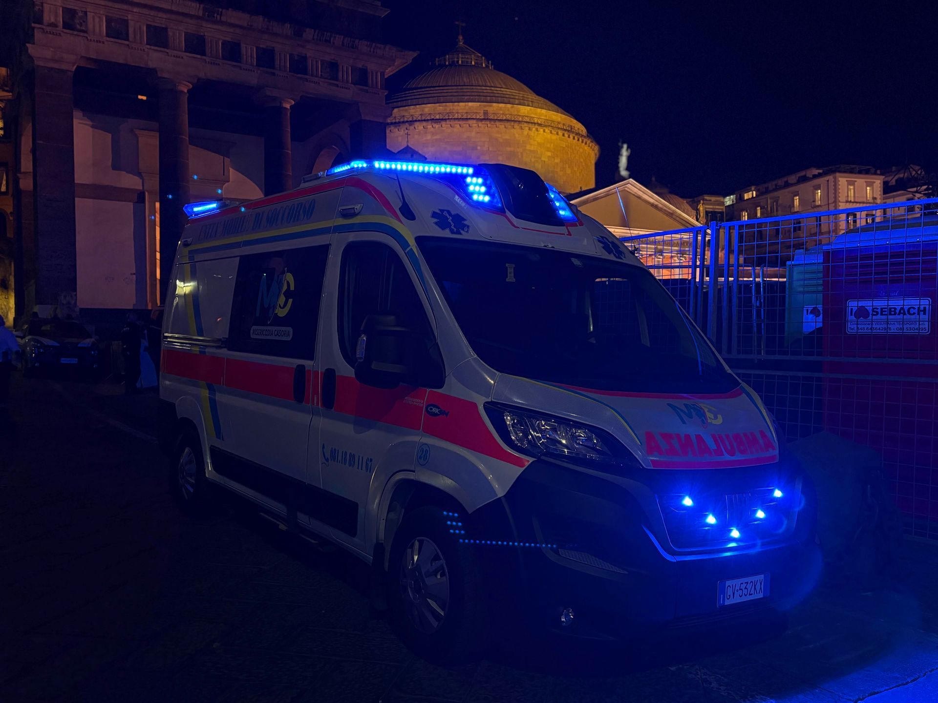Ambulanza con luci blu illuminate di notte, parcheggiata vicino a un edificio con colonne.