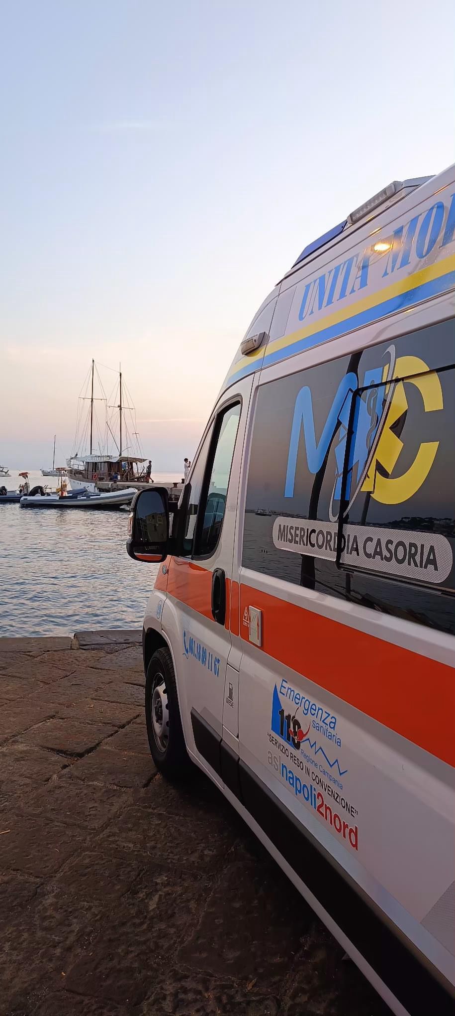 Ambulanza parcheggiata vicino all'acqua; barche e cielo sullo sfondo. Veicolo bianco con dettagli rossi e blu.