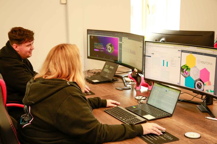 A man and a woman are sitting at a desk with laptops and monitors.