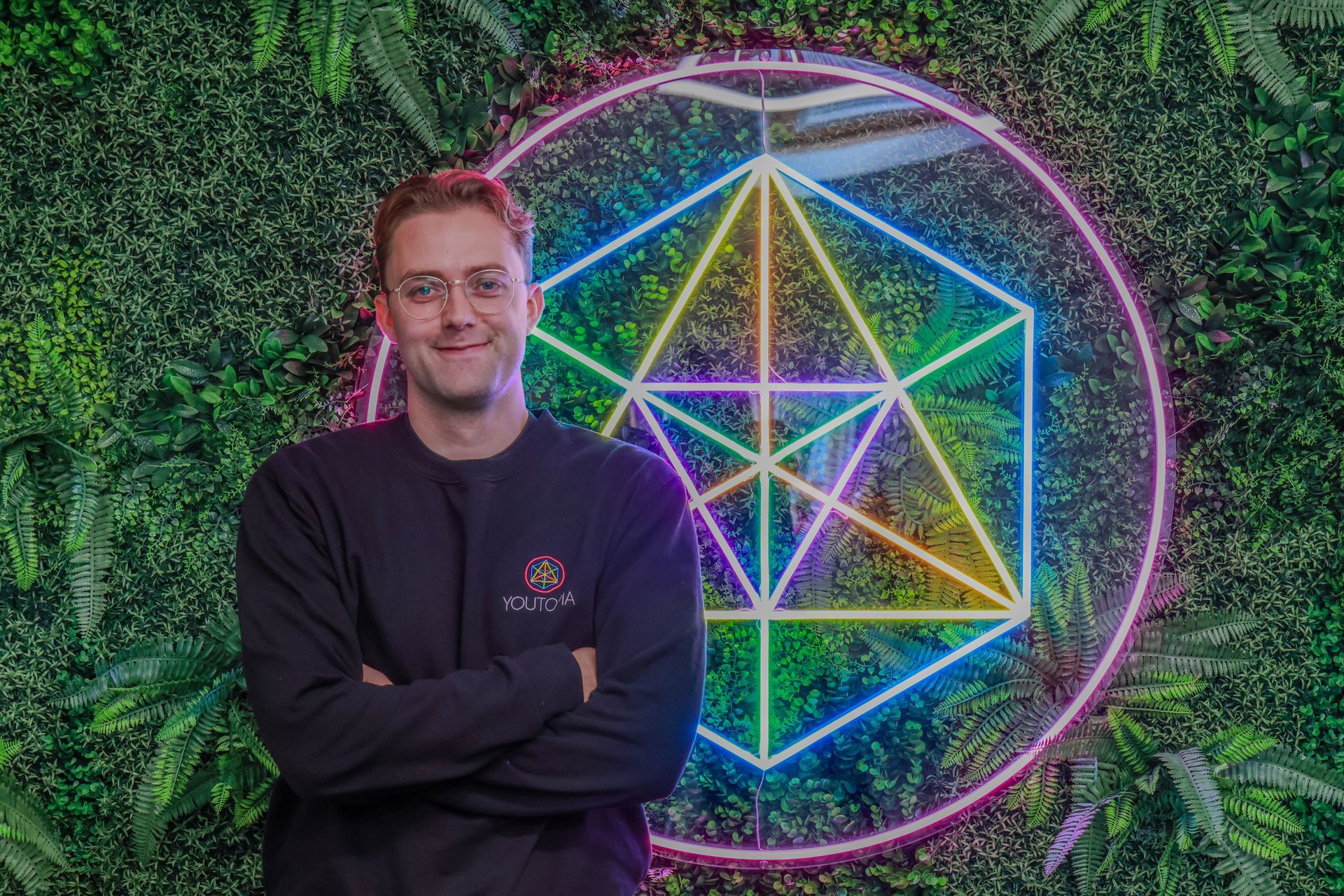 Man with glasses, arms crossed, smiles. Neon geometric shapes and greenery in background.