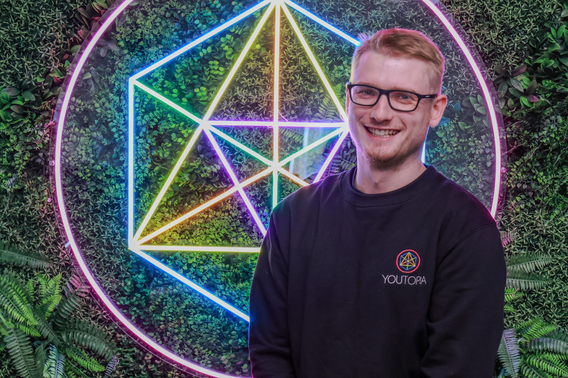Man with glasses smiling, standing in front of neon geometric shape on a green wall.