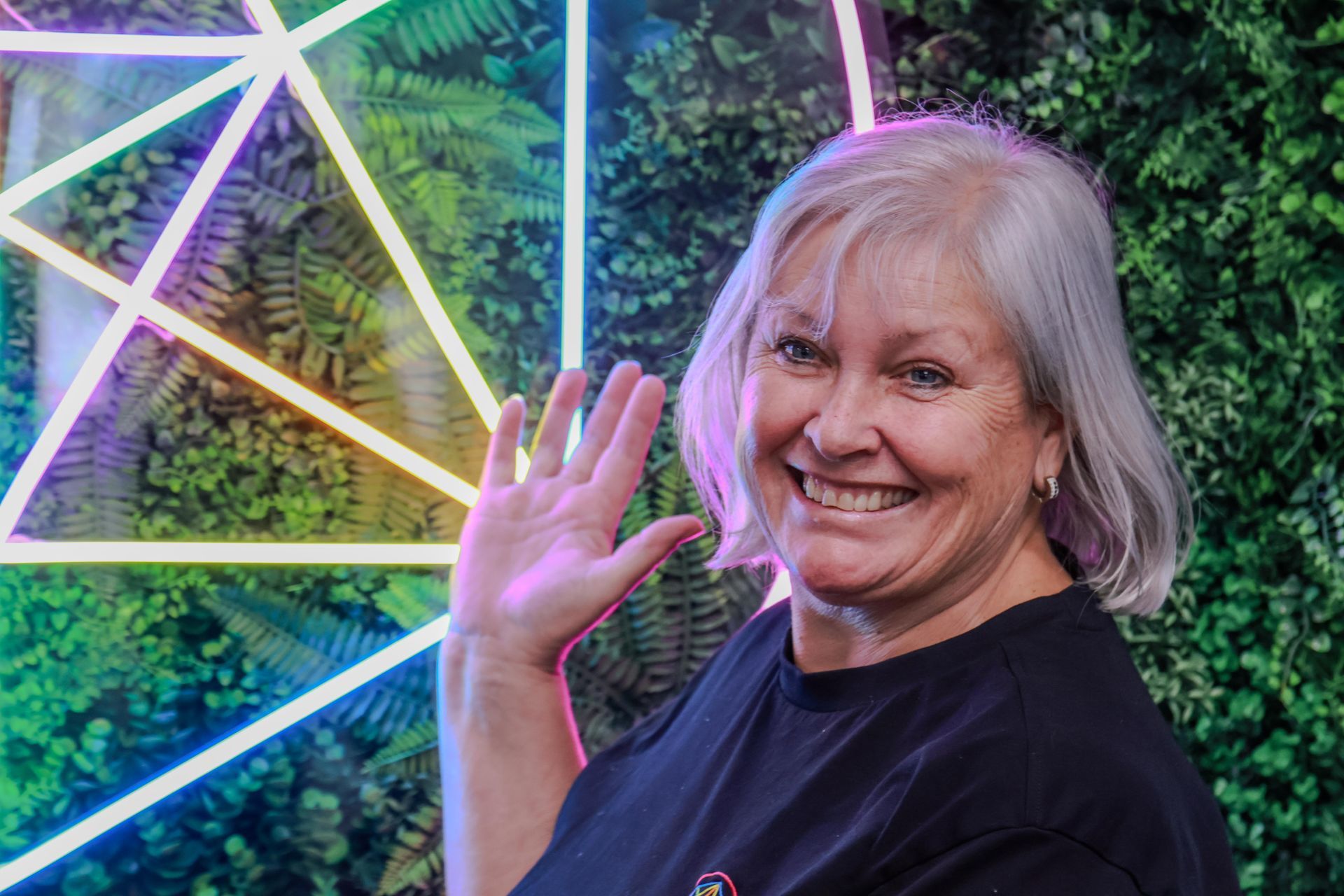 Woman with gray hair waves, smiling in front of green foliage and neon geometric shape.