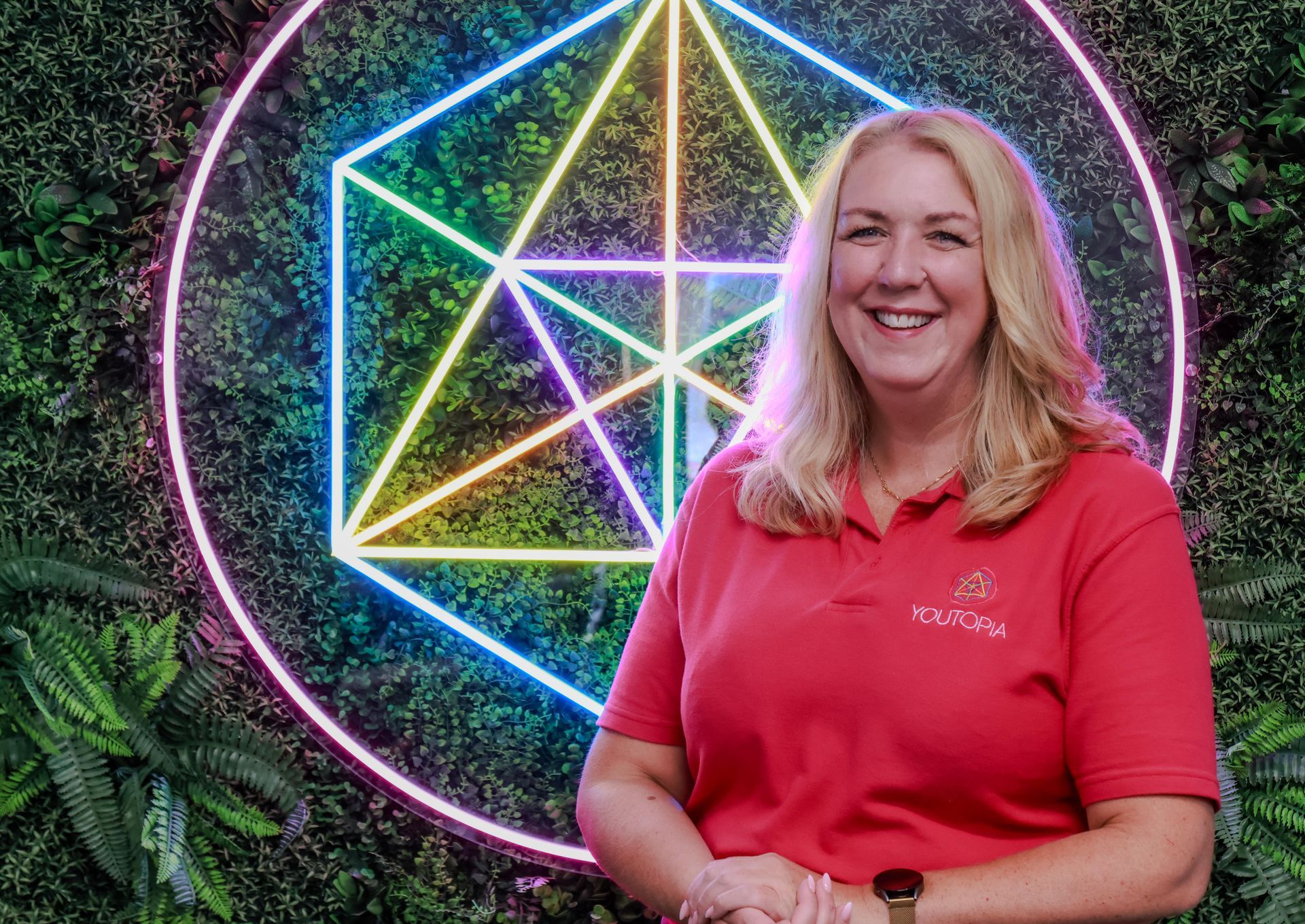 Woman in red shirt smiles in front of neon geometric shape on a green wall.