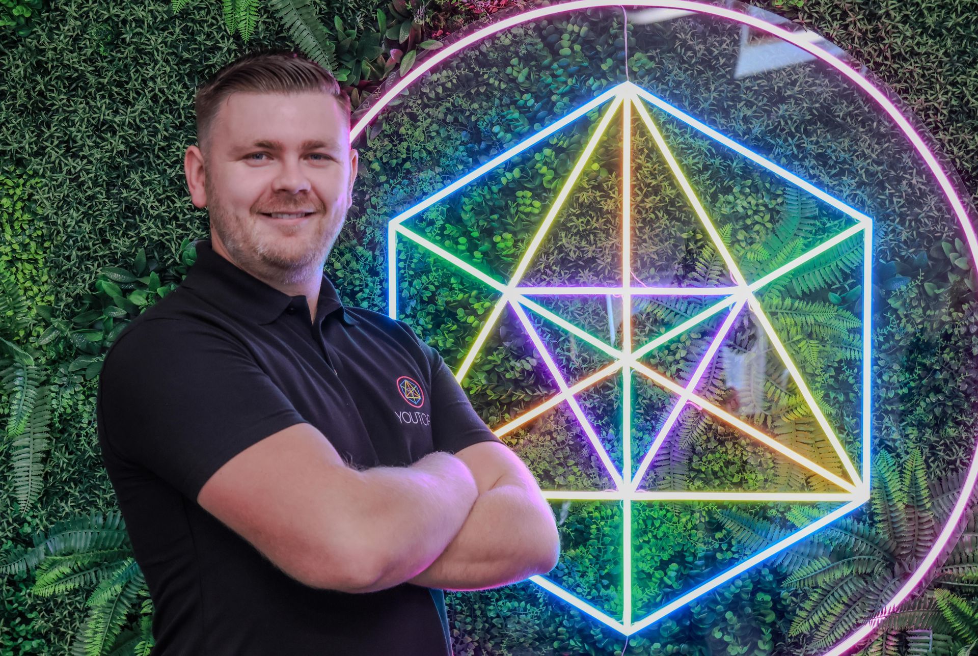 Man with arms crossed smiling in front of a neon geometric shape and green foliage.
